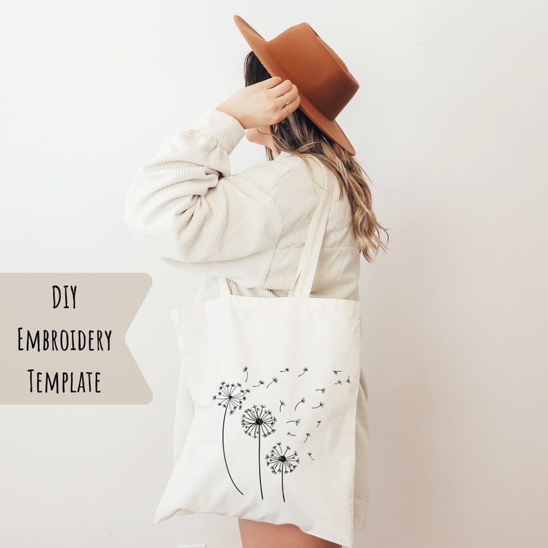 May include: Cream-colored tote bag featuring a black dandelion design with the text "DIY EMBROIDERY TEMPLATE". The bag is carried over the shoulder. The person in the image is wearing a brown hat and a cream-colored shirt.