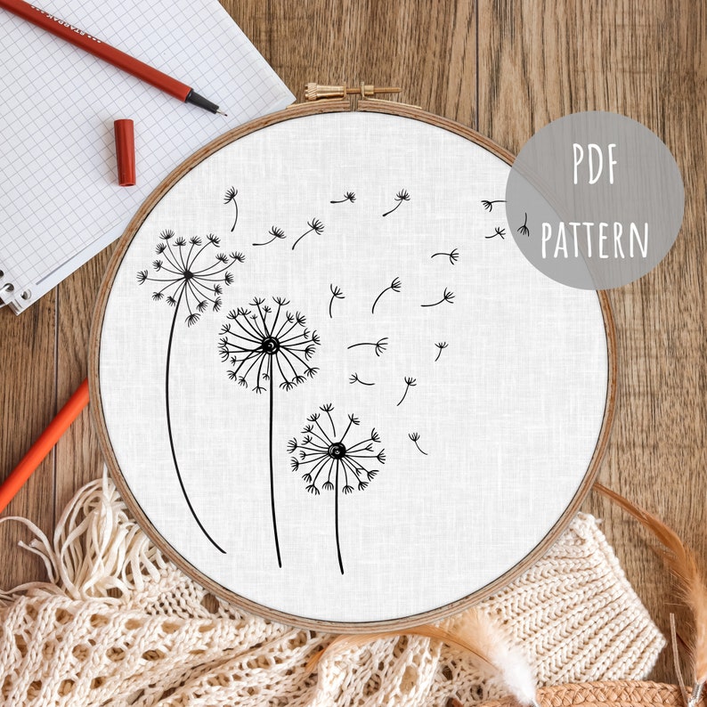 May include: A round embroidery hoop with a white fabric featuring a black line drawing of three dandelions with seeds blowing in the wind. The text "PDF PATTERN" is in a gray circle. A notebook and pencils are in the background.