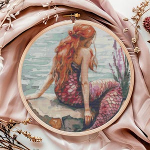 May include: A mermaid with long red hair sits on a rock by the ocean. Her tail is a vibrant pink and purple with scales. The water is a light blue with white waves.