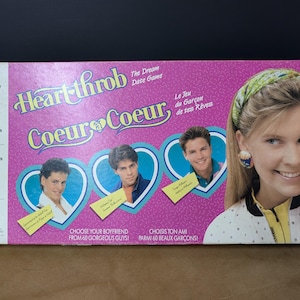 Op de afbeelding: Een vintage bordspeldoos met de titel "Heartthrob Coeur Coeur" in roze en blauw. De doos bevat afbeeldingen van jonge mannen en een vrouw. Tekst op de doos is onder meer "Kies je vriend uit 60 prachtige jongens!"