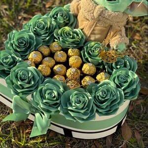 May include: A heart-shaped box filled with green silk roses and a variety of gold-wrapped chocolates. A small green plush toy with large ears sits on top of the arrangement.