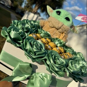 May include: A green and brown plush toy of a character from the Star Wars franchise, sitting in a heart-shaped box filled with green silk roses and gold chocolate candies.