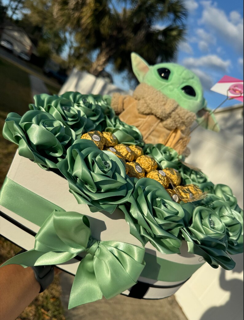May include: A green and brown plush toy of a character from the Star Wars franchise, sitting in a heart-shaped box filled with green silk roses and gold chocolate candies.