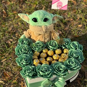 May include: A green plush toy character, resembling a baby, sits in a heart-shaped box filled with green roses and gold chocolate candies. A pink sign with the text "Fancy Gifts" is attached to the toy.