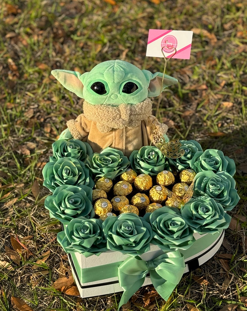 May include: A green plush toy character, resembling a baby, sits in a heart-shaped box filled with green roses and gold chocolate candies. A pink sign with the text "Fancy Gifts" is attached to the toy.