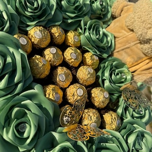 May include: A green and gold chocolate bouquet with a large arrangement of green satin roses surrounding a center of gold wrapped chocolates. A gold butterfly embellishment is on the right side of the bouquet.