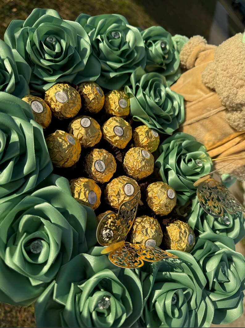 May include: A green and gold chocolate bouquet with a large arrangement of green satin roses surrounding a center of gold wrapped chocolates. A gold butterfly embellishment is on the right side of the bouquet.