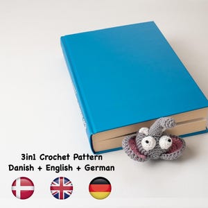 Pode incluir: Um livro azul com um marcador de elefante de crochê cinza. O livro tem o texto "3in1 Crochet Pattern Danish + English + German" na capa. As bandeiras da Dinamarca, do Reino Unido e da Alemanha são exibidas abaixo do texto.