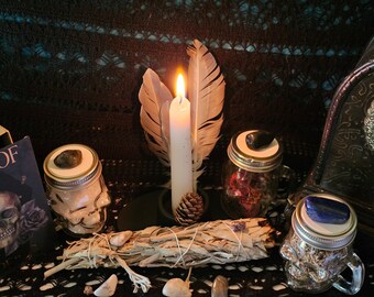 May include: A white candle burning with two feathers on either side, a bundle of sage, and three glass jars with crystals on top. The jars are decorated with skull designs.