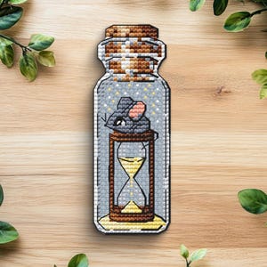 May include: A cross-stitch embroidery patch shaped like a glass bottle with a cork stopper. Inside the bottle, a gray mouse with pink ears sits atop a brown hourglass filled with yellow sand. The background inside the bottle is light gray.