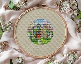 May include: A completed cross-stitch embroidery of a house with a red door, surrounded by a green lawn and pink flowering trees. The design is set within a wooden embroidery hoop, resting on a soft, cream-colored fabric.