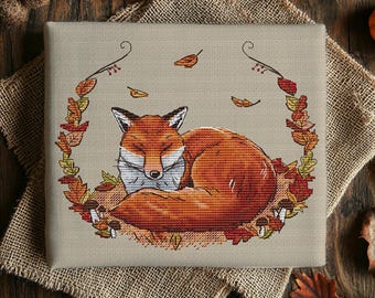 May include: A rectangular cross-stitch embroidery on beige fabric featuring a sleeping orange fox curled in a circle of autumn leaves and small mushrooms. The design includes delicate brown vines framing the fox with scattered falling leaves.