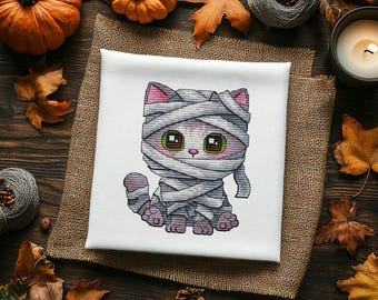 Puede incluir: Un cuadrado de tela blanca con un bordado de punto de cruz de un gatito envuelto en vendas de momia grises. El gatito tiene grandes ojos verdes, interior de las orejas rosa y una pequeña nariz rosa. El diseño está centrado en la tela.