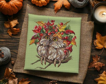 May include: A square cross-stitch embroidery on light green fabric featuring a brown owl perched on a branch. The owl is surrounded by a colorful wreath of autumn leaves in shades of red, orange, and yellow, accented with small red berries.