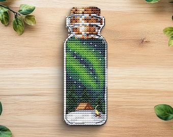 May include: A cross-stitch design of a jar containing a green and white aurora borealis scene. The jar has a brown cork top and a snowy base with a small tent and campfire. The background is a light wood grain.