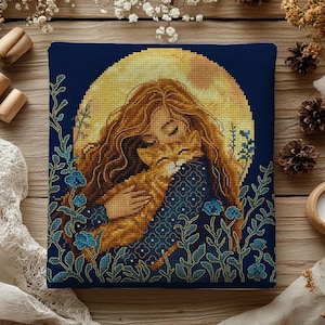Op de afbeelding: Kruissteekkunst met een vrouw die een kat omhelst, tegen een gele maan. De vrouw en kat zijn in bruin- en goudtinten, met een donkerblauwe achtergrond en bloemmotieven. Het werk is ingelijst.