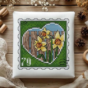 May include: A square white canvas featuring a cross-stitch design of a postage stamp. The stamp has a green border and a heart-shaped center containing yellow daffodils against a brown wooden fence background. The number 70 is in the corner.