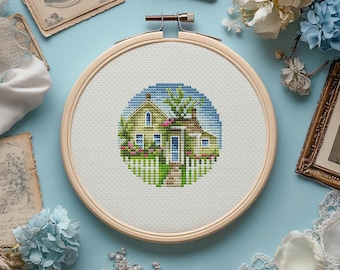 May include: A completed cross-stitch embroidery of a house with a white picket fence, surrounded by flowers and greenery, set within a wooden embroidery hoop. The house is yellow with a blue door and windows, and a blue sky background.