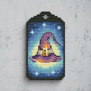 May include: A rectangular cross-stitch tag featuring a purple witch hat with a yellow buckle, centred against a large pale yellow moon and a dark blue starry night sky. The design is bordered by a black stitched frame with a hole at the top.