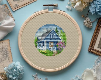 May include: A cross-stitch embroidery hoop featuring a blue house with a white porch and a gray roof, surrounded by green foliage and pink flowers. The embroidery is set against a beige fabric, framed by a wooden hoop.