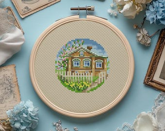 May include: A cross-stitch embroidery of a house with a white picket fence, trees, and a blue sky, set within a wooden embroidery hoop. The house is brown with blue windows and a green lawn. The hoop is set against a light blue background.