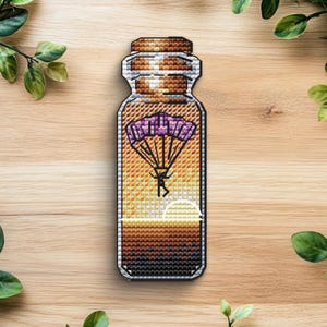 Puede incluir: Un parche de punto de cruz con forma de botella, que representa a un paracaidista bajo un paracaídas morado contra un fondo de puesta de sol. La botella tiene una tapa de corcho y está llena de capas de color.