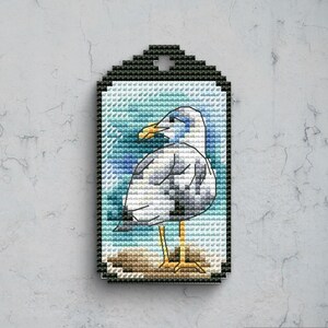 Könnte beinhalten: Ein rechteckiger Anhänger mit einem Kreuzstichmuster einer Möwe mit weißen und grauen Federn, einem gelben Schnabel und gelben Beinen. Der Vogel steht auf einer braunen Fläche vor einem blau-weißen Hintergrund, umrahmt von einem schwarzen Rand.
