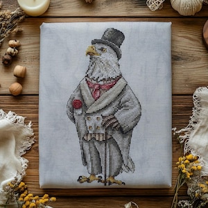 May include: A cross-stitch embroidery on light gray fabric featuring a bald eagle dressed in a formal gray tailcoat, a black top hat, a red cravat, and a red pocket square, while holding a thin black walking cane in its feathered wing.