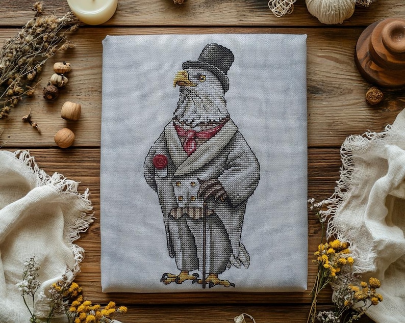 May include: A cross-stitch embroidery on light gray fabric featuring a bald eagle dressed in a formal gray tailcoat, a black top hat, a red cravat, and a red pocket square, while holding a thin black walking cane in its feathered wing.