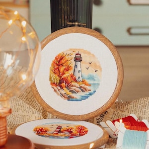 Puede incluir: Una bordadura de punto de cruz enmarcada que representa un faro en una costa rocosa con árboles de otoño en el fondo. La escena se desarrolla en un cielo azul con algunas aves volando por encima.