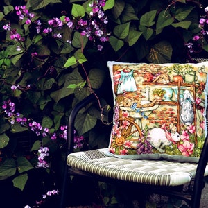 May include: A decorative square pillow with a whimsical illustration of rabbits, flowers, and a brick wall scene. The pillow is placed on a black metal chair with a woven seat, set against a backdrop of lush green foliage and purple flowers.