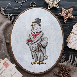 May include: An oval embroidery hoop containing a cross-stitch design of a bald eagle dressed in a formal gray tailcoat, a top hat, a red cravat, and a red rose boutonniere, holding a walking cane against a light gray fabric background.