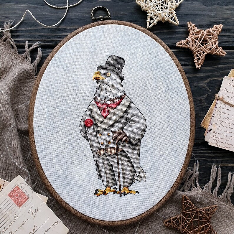 May include: An oval embroidery hoop containing a cross-stitch design of a bald eagle dressed in a formal gray tailcoat, a top hat, a red cravat, and a red rose boutonniere, holding a walking cane against a light gray fabric background.