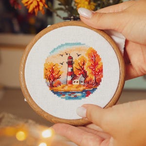 Puede incluir: Un aro de bordado blanco con un diseño de punto de cruz de un faro rojo y blanco con una pequeña casa a su lado. El faro está rodeado de árboles con hojas naranjas, amarillas y rojas. El fondo es un cielo azul con nubes blancas.