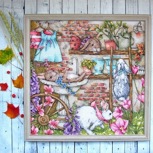 May include: Cross-stitch artwork featuring a whimsical scene with several rabbits in a garden setting. The artwork includes a brick wall, flowers, and a dress hanging on a clothesline. The rabbits are depicted in various poses, adding to the charm of the piece.