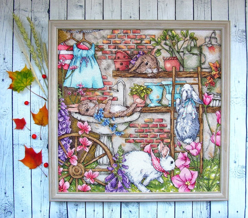 May include: Cross-stitch artwork featuring a whimsical scene with several rabbits in a garden setting. The artwork includes a brick wall, flowers, and a dress hanging on a clothesline. The rabbits are depicted in various poses, adding to the charm of the piece.