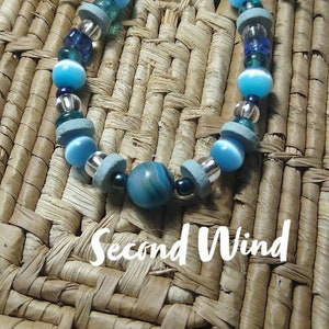 Può includere: Un braccialetto di perline blu e bianche con una perla di agata blu e bianca al centro. Il braccialetto è su una superficie intrecciata e il testo "Second Wind" è visibile sotto il braccialetto.