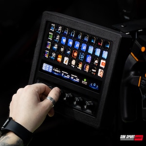 May include: A black control panel with multiple illuminated icons and buttons. The panel has a series of small screens displaying various app icons. A hand is adjusting a knob on the panel. The panel is part of a racing simulator setup.