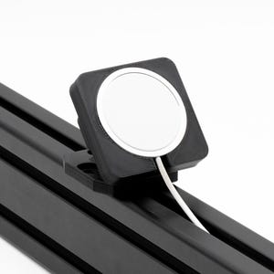 Puede incluir: Un soporte de carga cuadrado negro con una base de carga circular blanca y un cable blanco. El soporte está montado sobre un riel metálico negro. El texto "SIM SPORT GADGET" está en la esquina superior derecha.