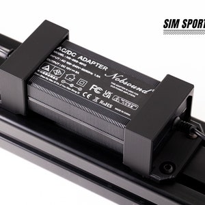 Könnte beinhalten: Ein schwarzer AC/DC-Adapter mit dem Text "AC/DC Adapter" und "Nobsound" darauf gedruckt. Der Adapter hat ein schwarzes Netzkabel und ist an einer schwarzen Metalloberfläche befestigt. Der Text auf dem Adapter lautet: "Modell: NS-100-0012", "Eingang: AC 100-240V~50/60Hz 1.8A", "Ausgang: DC 12V 5A", "Dieser Adapter wird für unseren Leistungsverstärker verwendet."