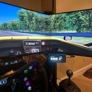 Puede incluir: Un simulador de carreras con un volante negro, una palanca de cambios negra y dos monitores grandes que muestran un juego de carreras. Los monitores están montados en un soporte negro. El volante tiene varios botones y una pantalla digital. La palanca de cambios tiene una base negra y una perilla plateada. Los monitores muestran un coche de carreras amarillo conduciendo en una pista con árboles a ambos lados.