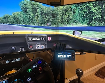 May include: A racing simulator setup with a black steering wheel, a black gear shifter, and two large monitors displaying a racing game. The monitors are mounted on a black stand. The steering wheel has a variety of buttons and a digital display. The gear shifter has a black base and a silver knob. The monitors display a yellow race car driving on a track with trees on either side.