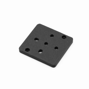 May include: A black, square metal plate with rounded corners. The plate has five circular holes and two hexagonal holes. The surface has a slightly textured appearance. The plate is designed for mounting or connecting components.