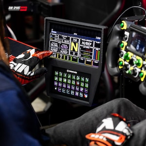 Puede incluir: Una pantalla digital negra que muestra datos de carreras, incluyendo RPM, combustible e información de vuelta. Debajo, un Stream Deck con múltiples iconos coloridos. Parte de una configuración de simulador de carreras.