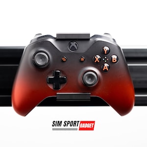 May include: A black and red Xbox controller with orange buttons is mounted on a black wall mount. The controller has a gradient fade from black to red. The wall mount is labeled "SIM SPORT GADGET".