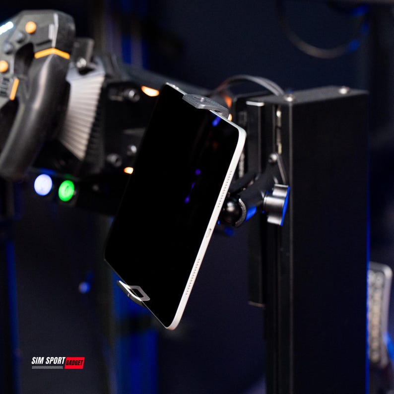 May include: A black tablet with a white border is mounted on a black metal stand. The stand is part of a racing simulator setup, with a steering wheel and other controls visible in the background. The text "SIM SPORT GADGET" is at the bottom.