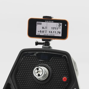 May include: A black and orange digital display device with a screen showing speed, RPM, and lap time data. The device is mounted on a black textured surface. The screen displays the text "SPD 0 km/h", "1711 RPM", and "13.11.78".