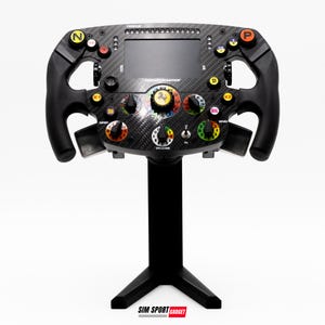 May include: A black and carbon fiber racing wheel with multiple colored buttons, dials, and a small screen. The wheel is mounted on a black stand. The text "THRUSTMASTER" is visible on the screen. The Ferrari logo is in the center.