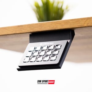 Może przedstawiać: Czarna klawiatura montowana na biurku z srebrnym panelem przycisków z etykietą "Sim Racing Deck". Klawiatura jest przymocowana do drewnianego biurka.