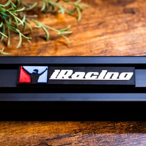 May include: Black rectangular object with the text "iRacing" in white. The logo features a red, white, and blue design. The object is set against a wooden background with a hint of greenery.
